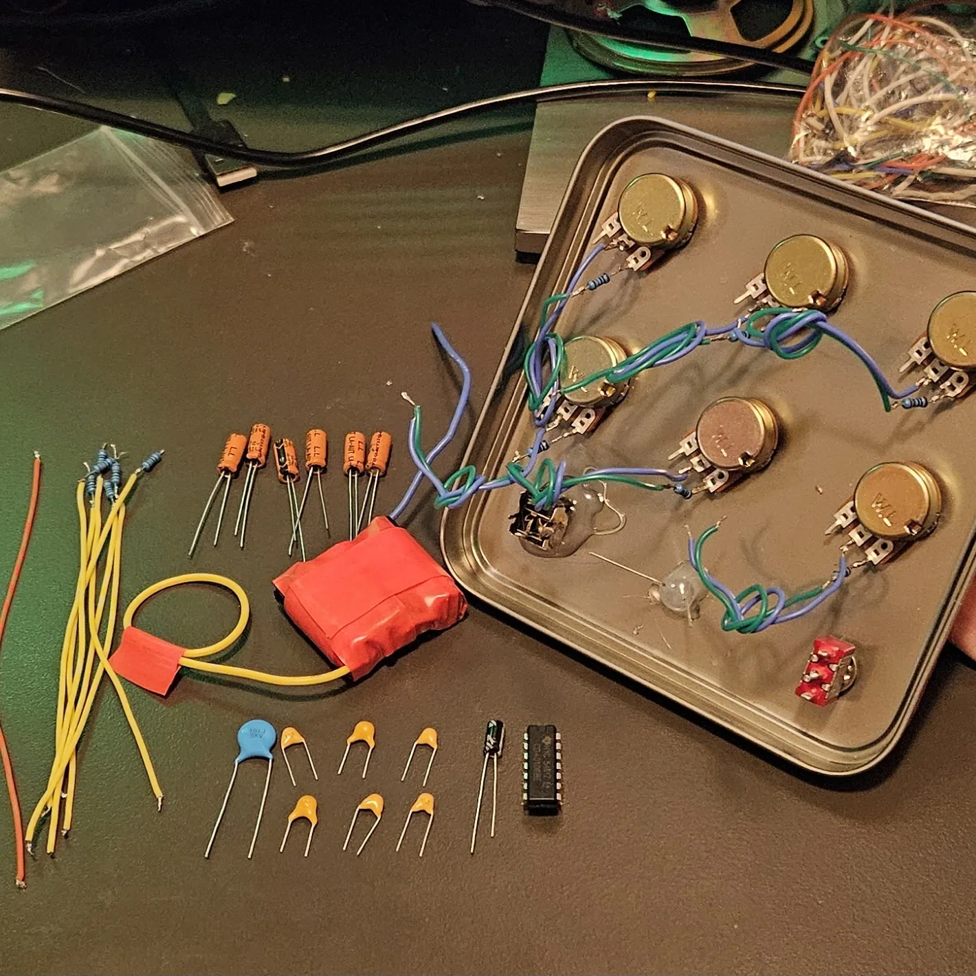 Everything for the build laid out on a workbench. On the left, lengths of orange and yellow hookup wire, a few small resistors, a row of orange electrolytic capacitors, and a battery pack wrapped in red tape. Along the bottom, the ceramic voice capacitors - one large blue disc beside a group of small yellow ones - an LED, and the black CD40106 chip. On the right sits the square tin with all six potentiometers already mounted through the lid and wired together with blue and green pairs, plus a red toggle switch and the output jack.