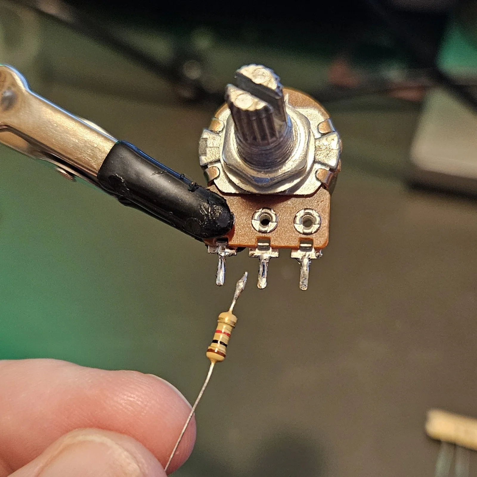 A potentiometer held in a helping-hands clip with one of its three solder lugs freshly tinned, and a colour-banded resistor held just below it with its own lead tinned, ready to be joined.