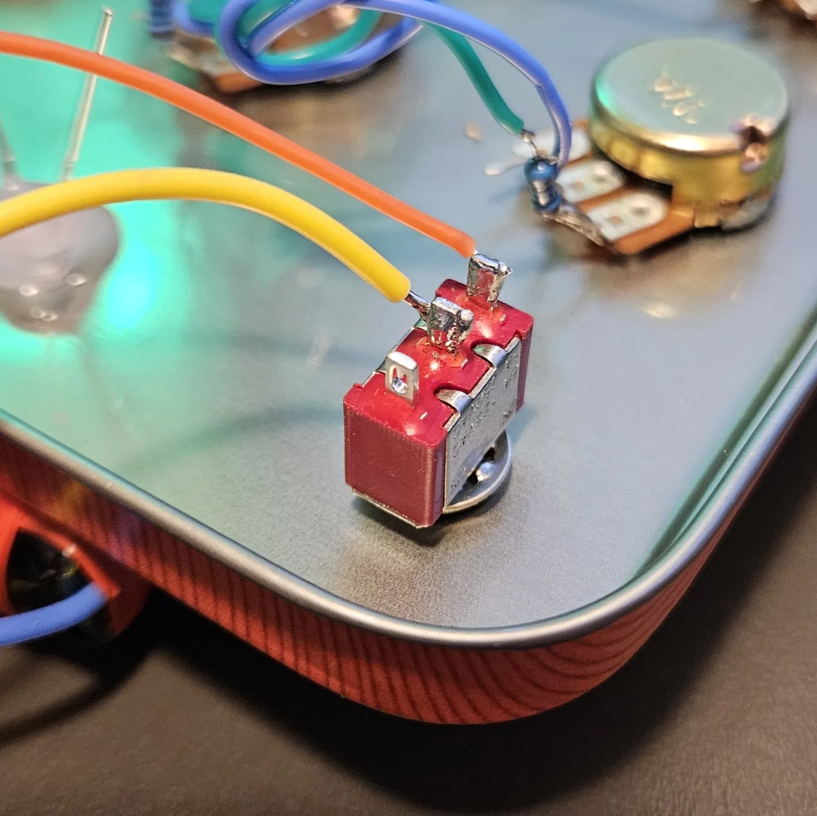 A red toggle switch mounted through the corner of the tin lid, with an orange and a yellow wire already soldered to its two lugs. A potentiometer with its own resistor and wires sits nearby on the panel.