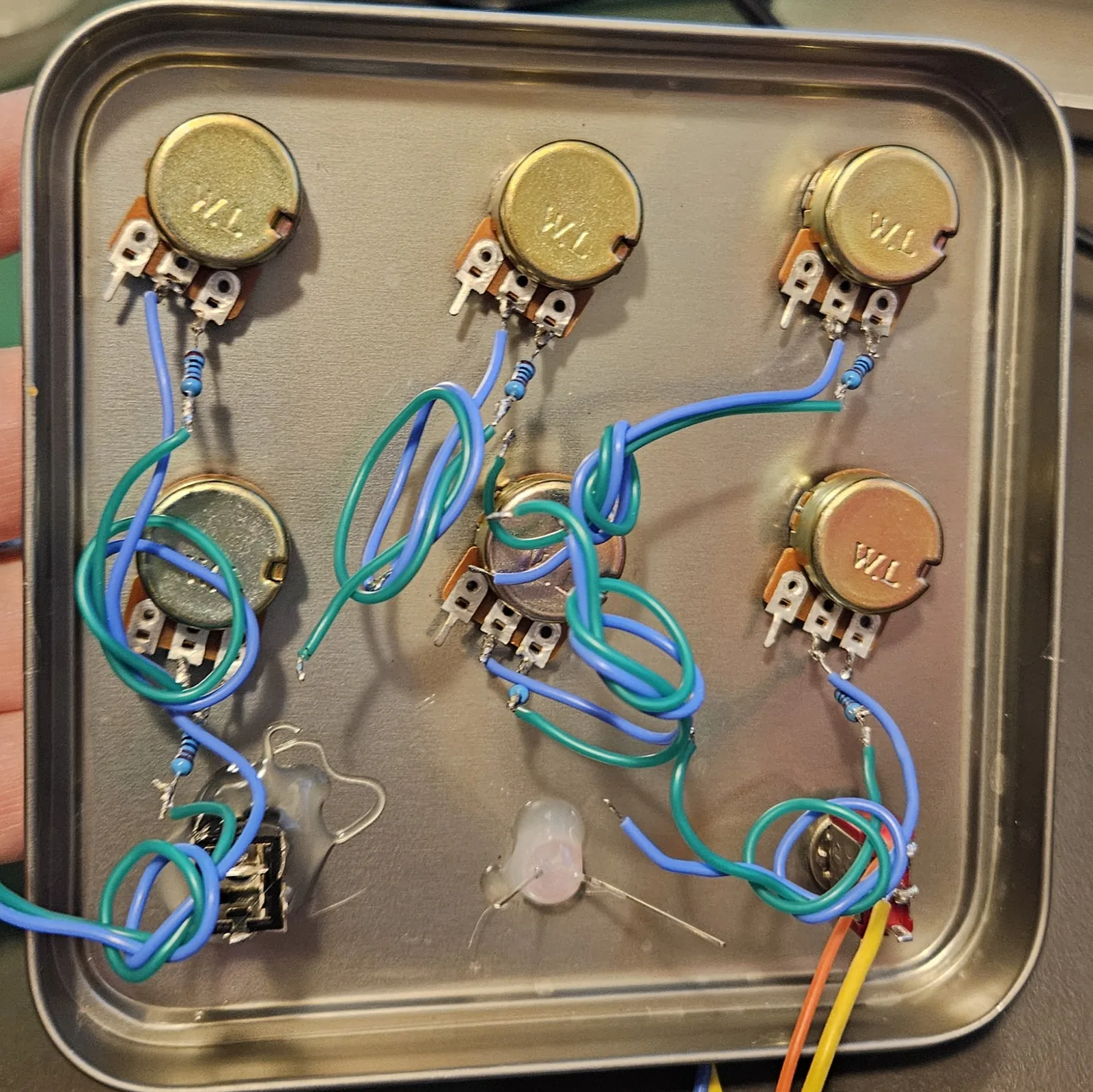 The inside of the tin lid fully populated: six potentiometers mounted through the panel in two rows of three, each with a small resistor soldered to one lug and blue and green wires running from them, plus an LED and a toggle switch mounted at the bottom and an output jack in the corner with orange and yellow leads.