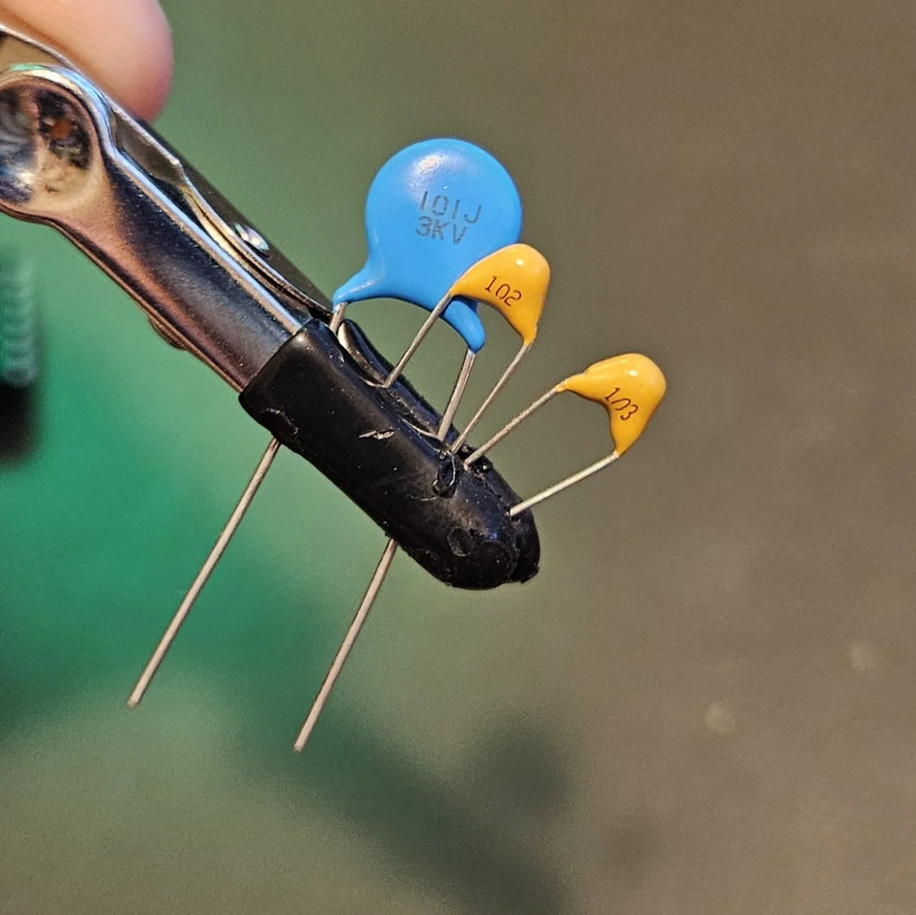 Three ceramic capacitors held side by side: a blue disc printed 101J 3KV and two smaller yellow ones printed 102 and 103.