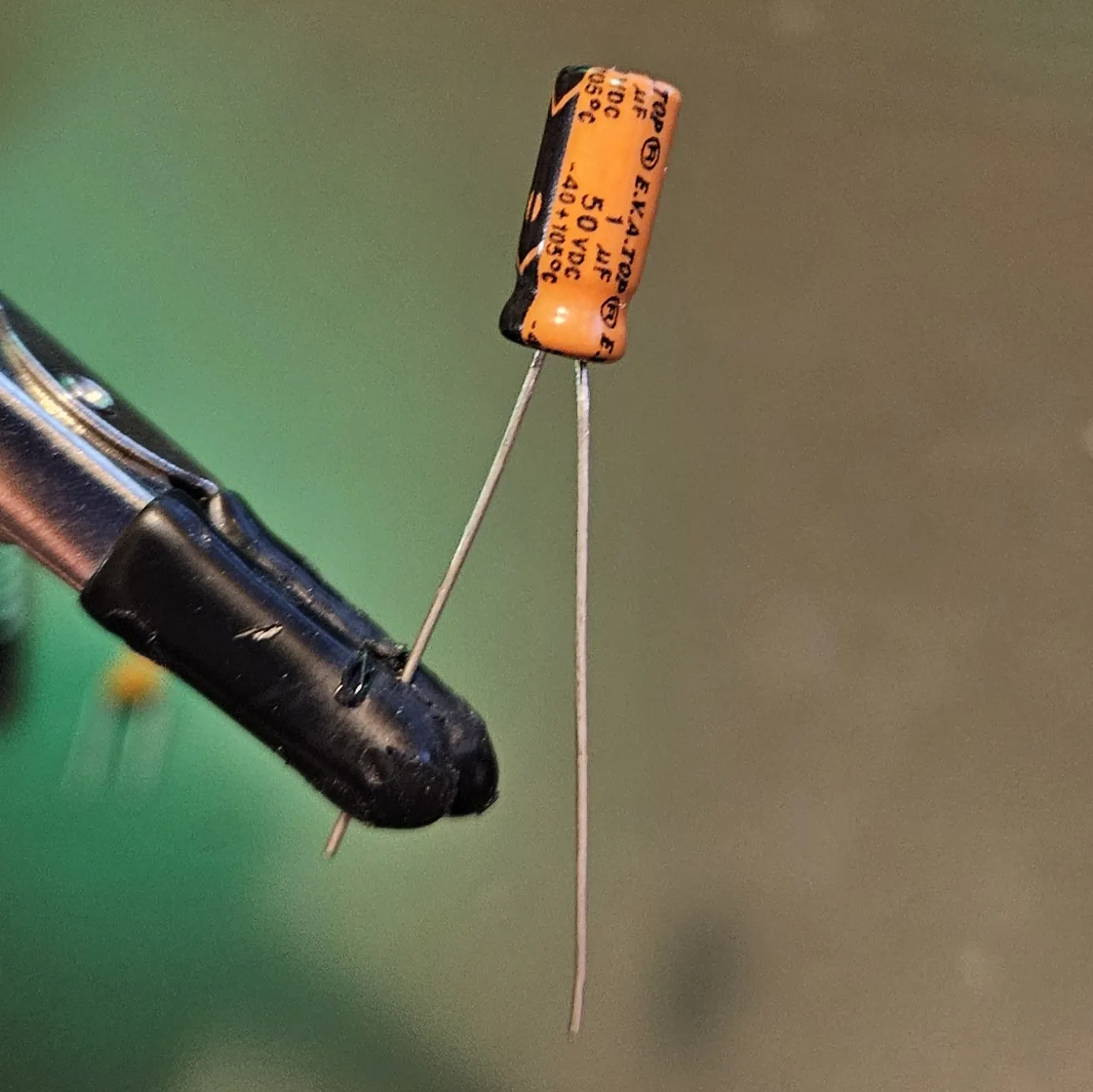 An orange cylindrical electrolytic capacitor held in a clip, printed 1 uF 50 VDC, with two long silver legs.