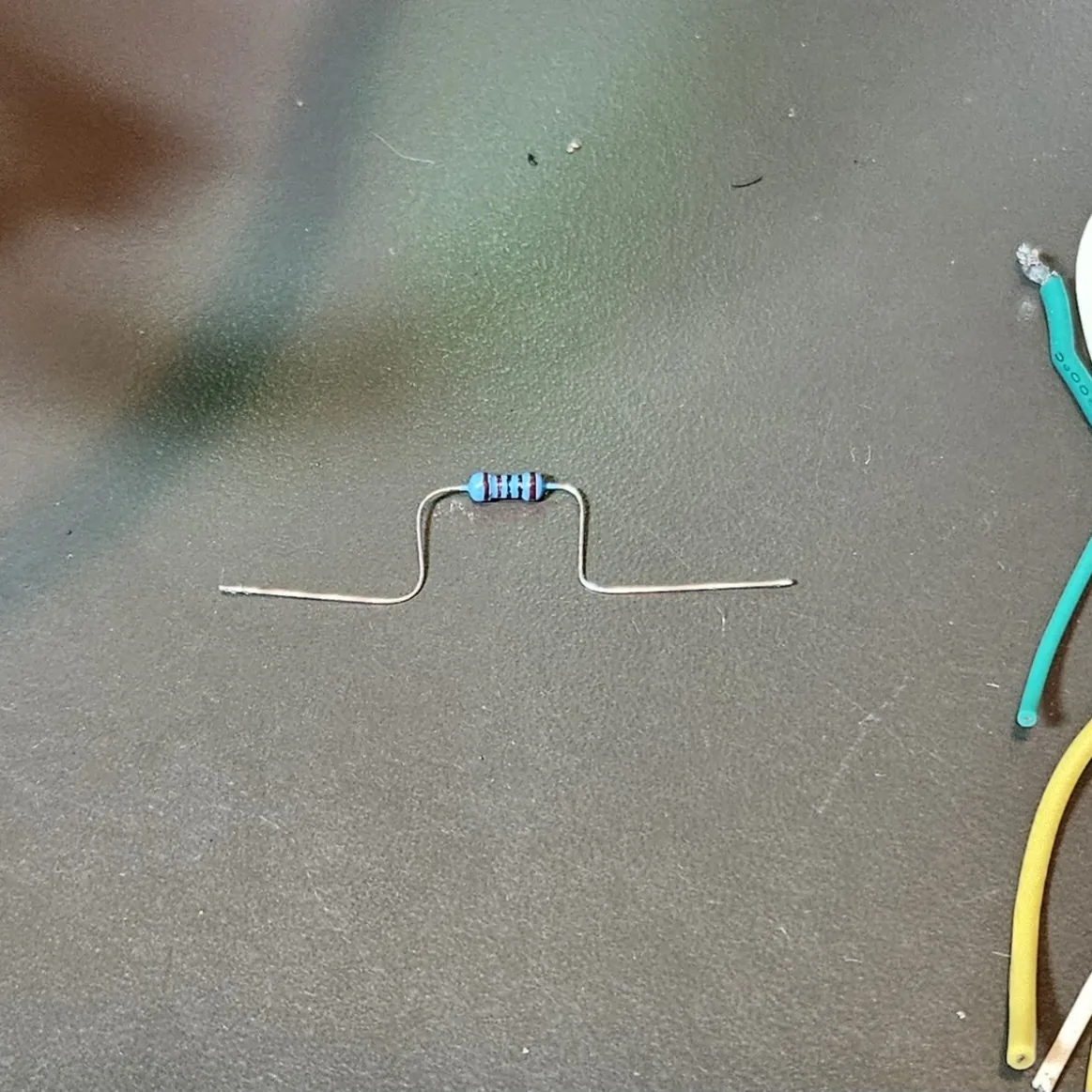 A blue metal-film 1K resistor lying on the mat with its leads pre-bent into a flat-topped staple shape, ready to drop onto two pins.