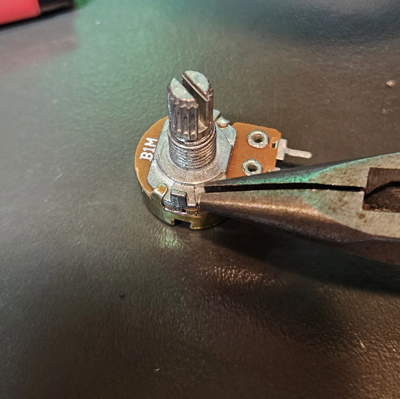 The B1M pitch potentiometer, its back label showing, with pliers gripping the small anti-rotation tab on the side of its bushing.
