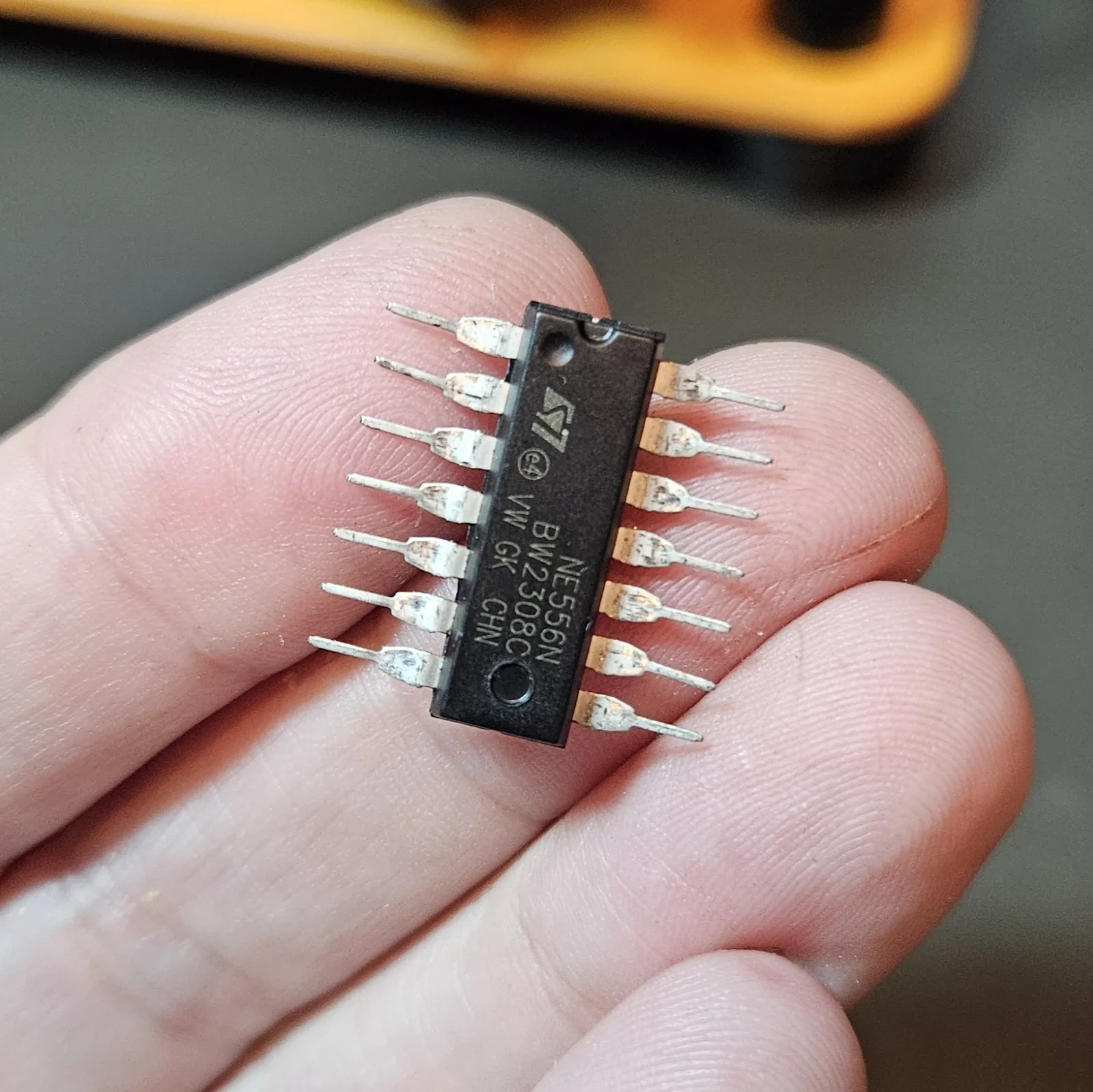 The NE556 held in fingers with its legs splayed straight out to both sides, the ST 'NE556N BW2308C' marking, the pin-1 notch, and the pin-1 dimple all visible.