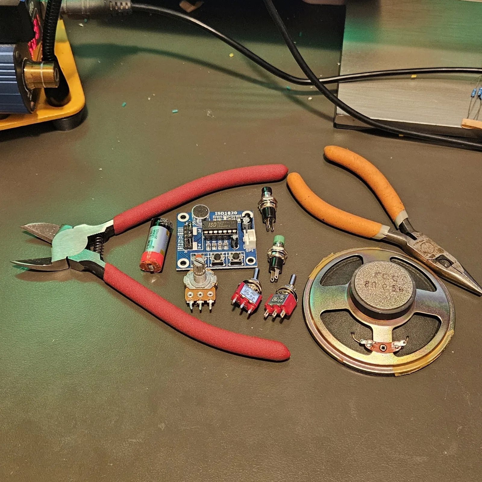 The parts for the build laid out on a workbench: red-handled flush cutters on the left, a small cylindrical lithium cell marked 3.7V, the blue ISD1820 module with its J1 header silkscreened VCC GND FT P-L P-E REC, a potentiometer, two red mini toggle switches, two green panel-mount momentary push buttons, a large gold-framed round speaker, and orange-handled needle-nose pliers. A soldering station and spool of solder sit at the top left.