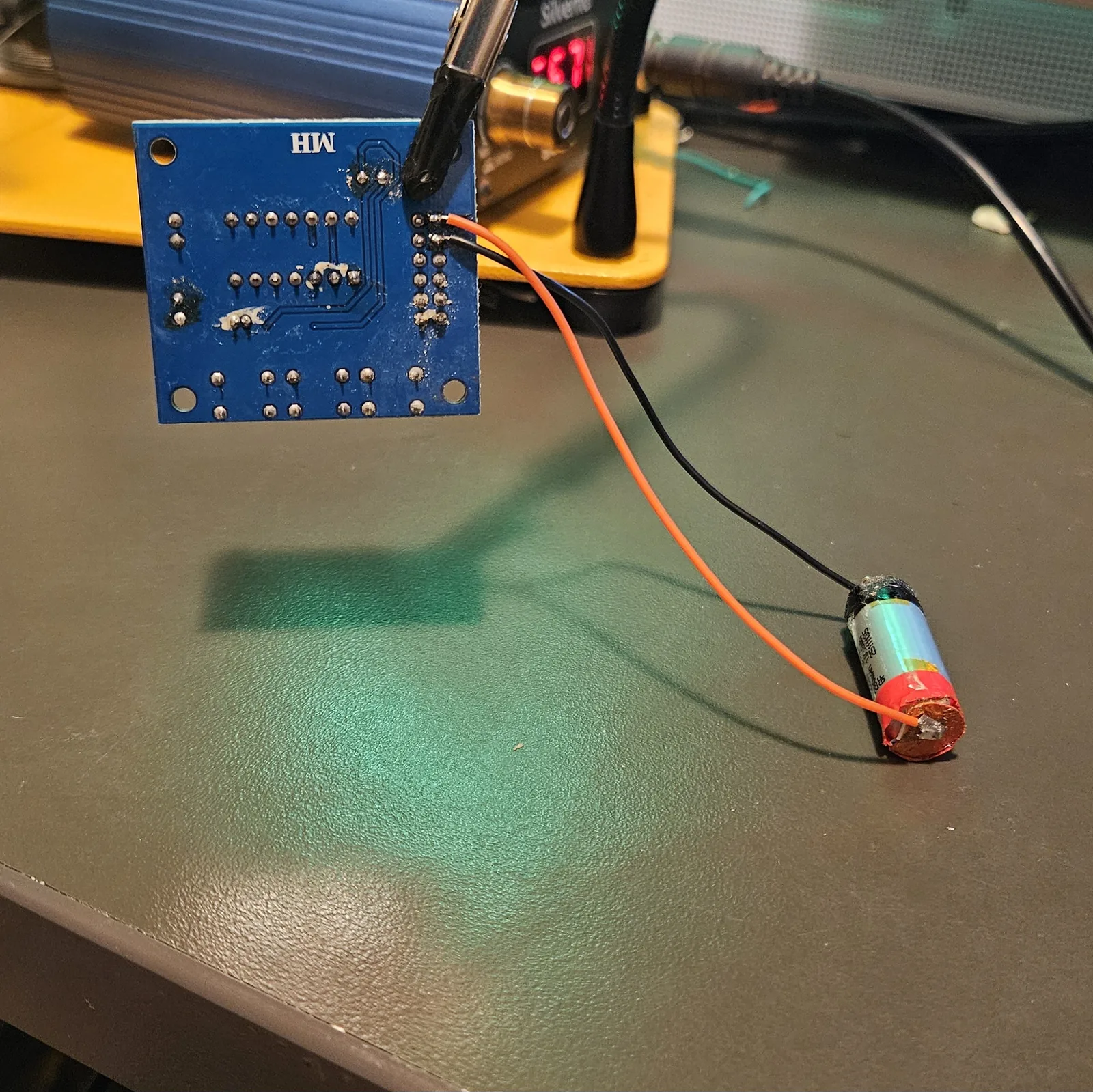 The module suspended in the helping-hands clip with its orange and black leads running out across the bench to a small cylindrical lithium cell.