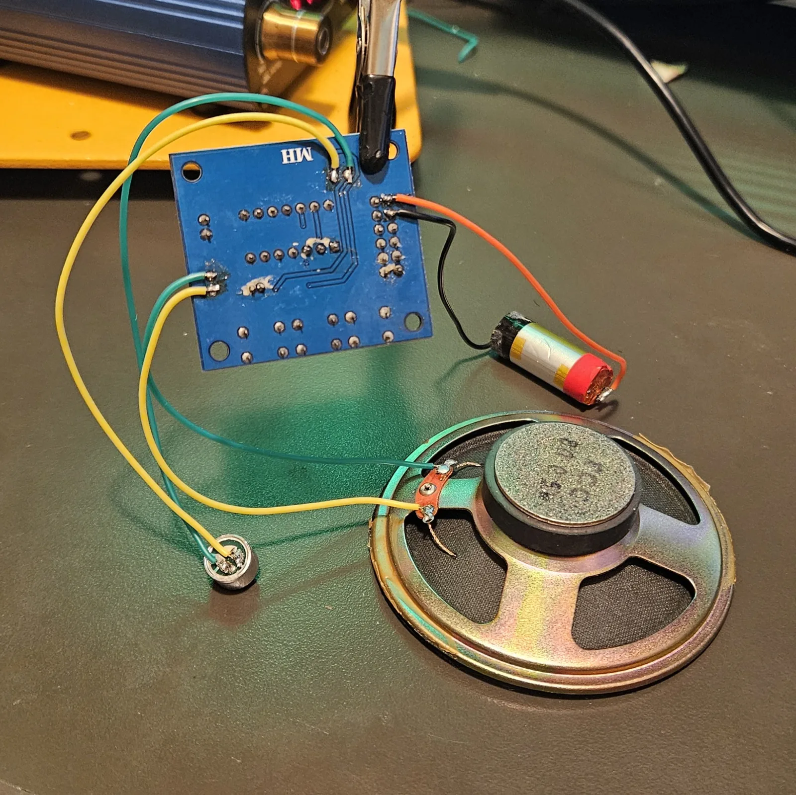The module on the bench with green and yellow wire pairs running out to two components: the salvaged electret microphone capsule on the left, now on flying leads, and the large gold-framed speaker on the right. Orange and black leads run to the lithium cell behind.