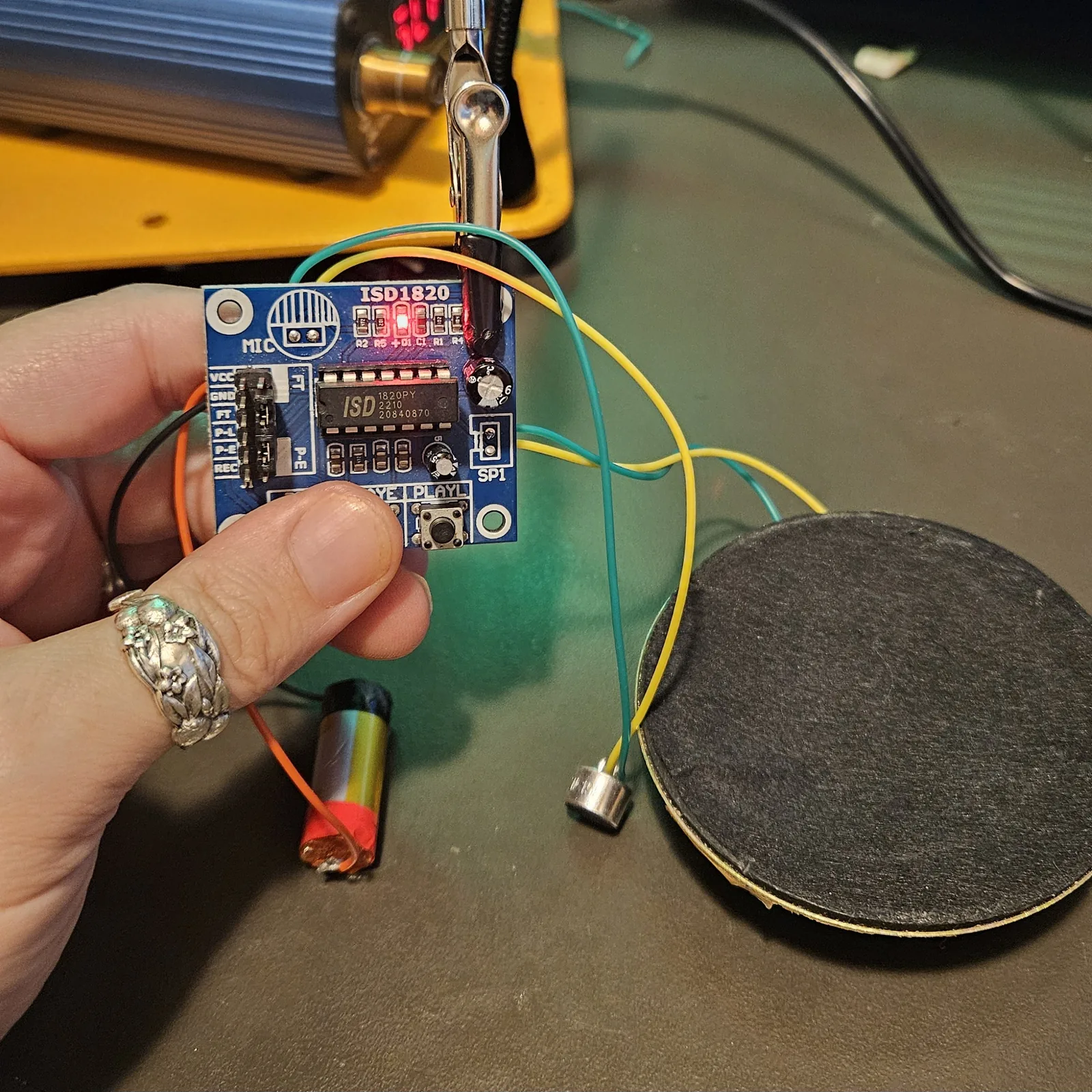 The finished wiring held up in one hand: the module with its D1 LED lit, the relocated electret microphone and the large speaker both connected on green and yellow leads, and the lithium cell hanging below.