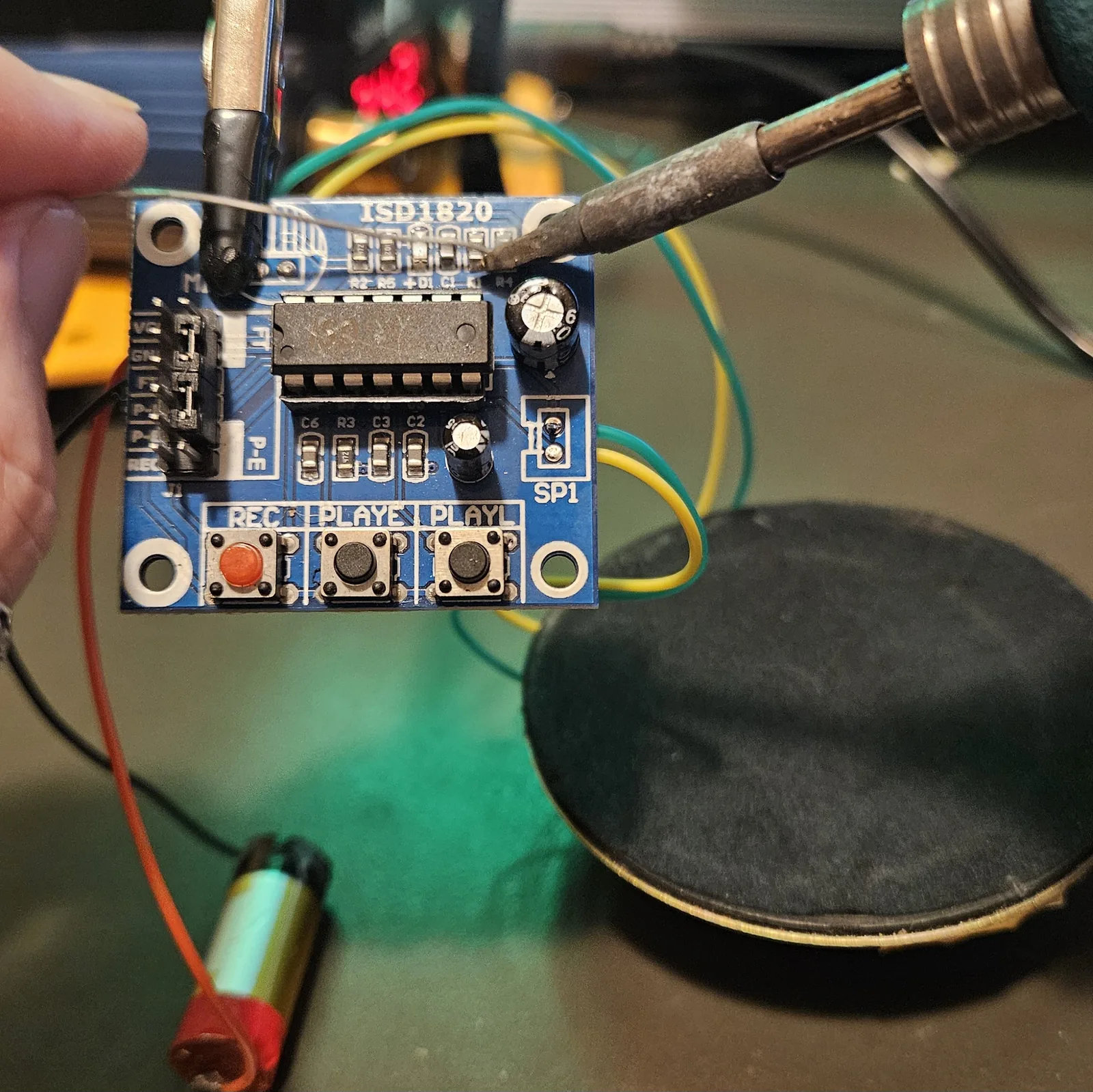A soldering iron tip pressed against the row of small surface-mount components along the top edge of the module, at the position marked R4, while the board is held steady in a helping-hands clip.