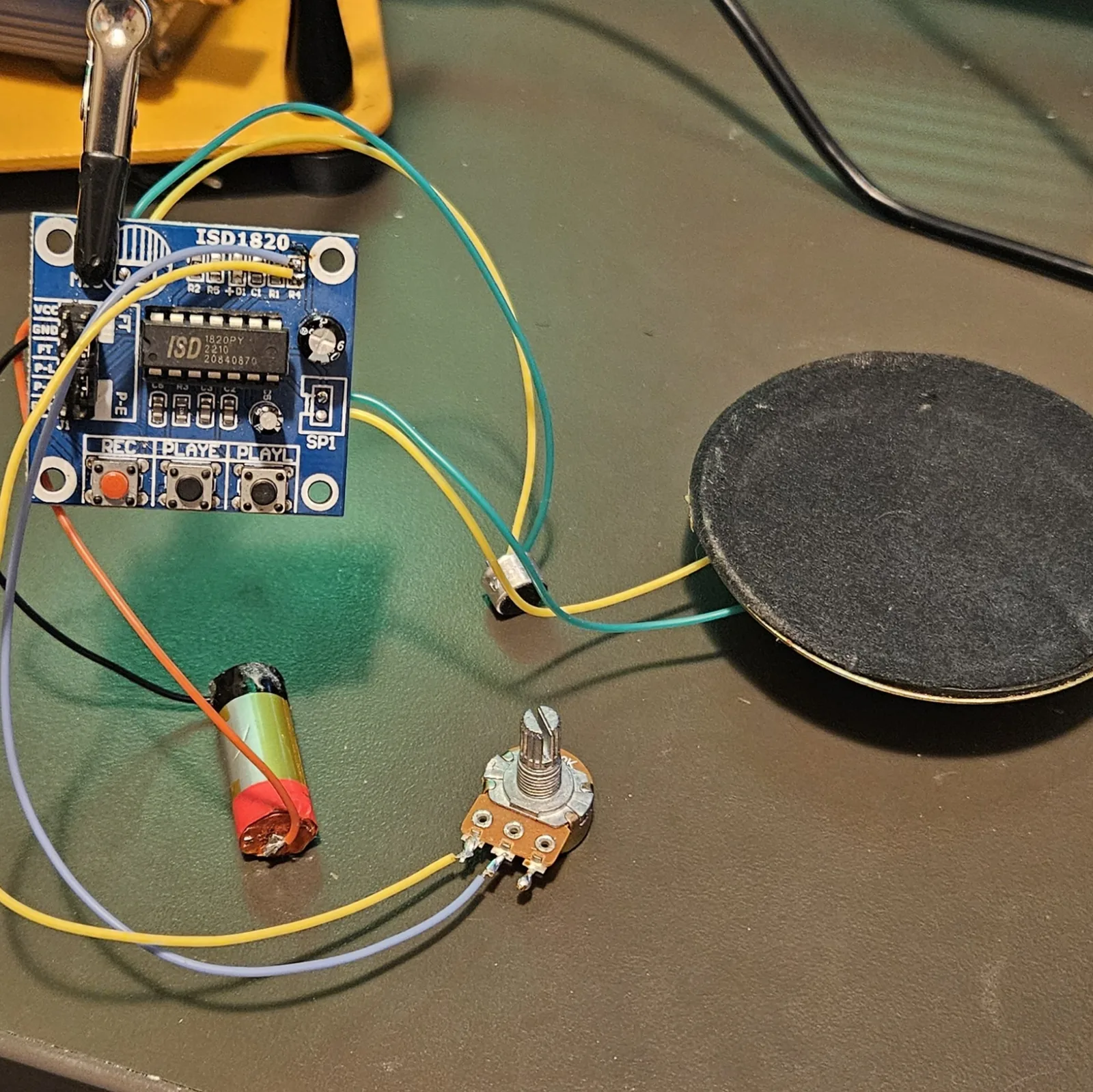 The whole assembly laid out on the bench: the module with its yellow and grey pitch-pot leads running to the potentiometer at the bottom, the electret mic and speaker on green and yellow leads, and the lithium cell alongside.