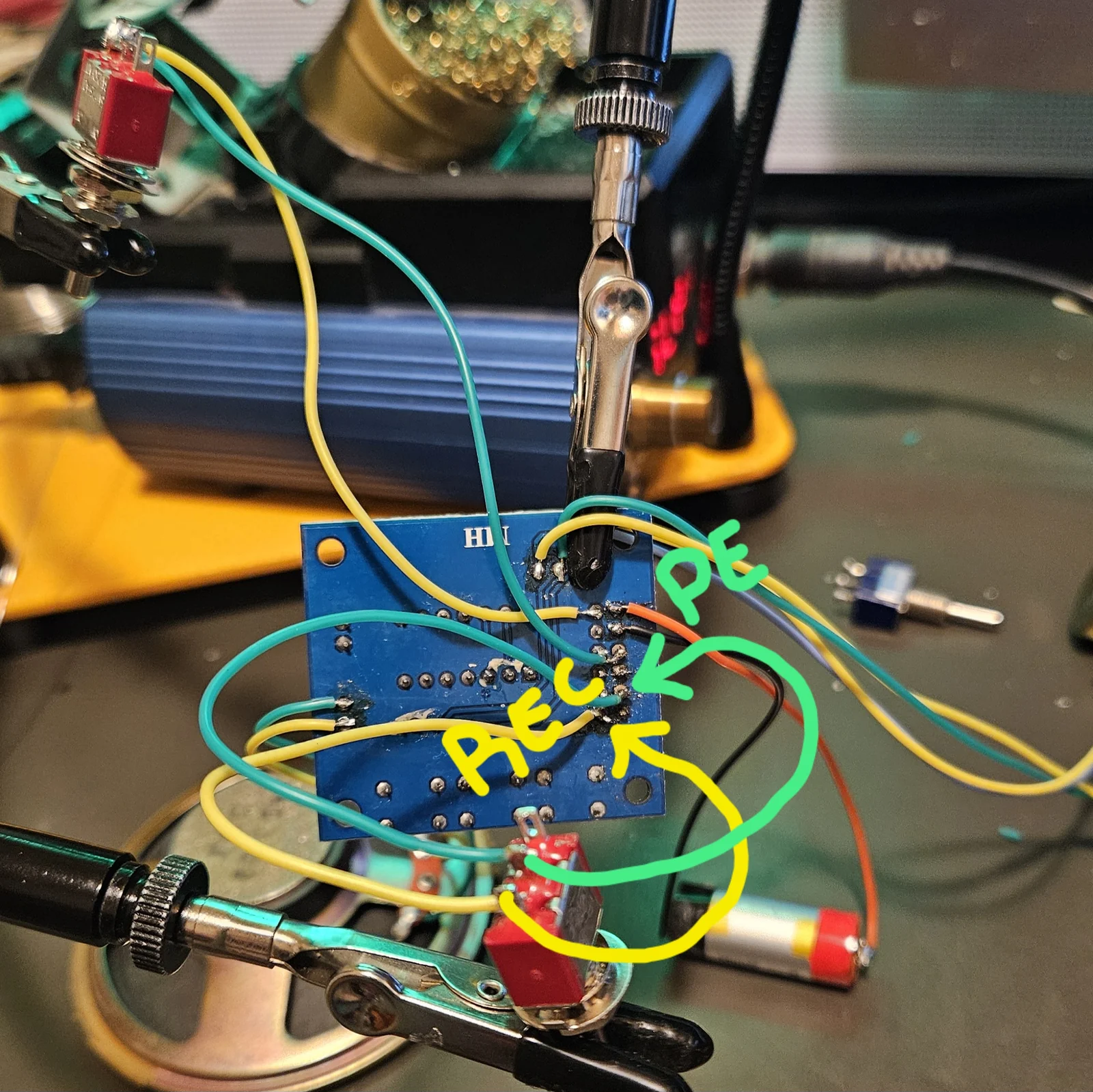 The underside of the ISD1820 module wired for the working build, held in helping hands, with hand-drawn annotations added. A green arrow labelled P-E points to a pad on the right-hand pad row, and the teal wire leaving it is traced in green down to a red toggle switch at the bottom. A yellow arrow labelled REC points to an adjacent pad, and its yellow wire is traced down to the same switch area. Orange and black power leads run off to a cylindrical lithium cell on the right.