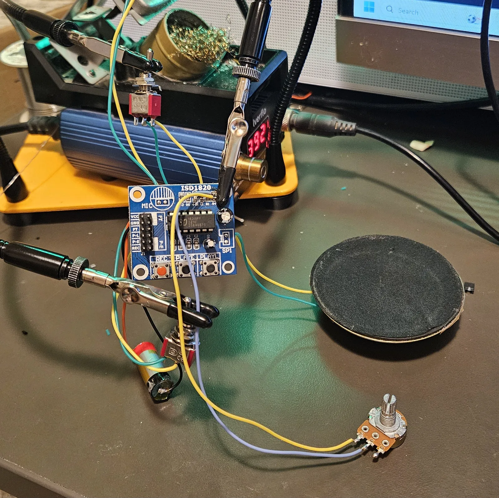 The complete build spread across the workbench: the ISD1820 module clipped in helping hands with its J1 header labels visible, two red toggle switches wired in, the lithium cell, the relocated electret microphone, the large speaker, and the pitch potentiometer, all connected with coloured hookup wire.
