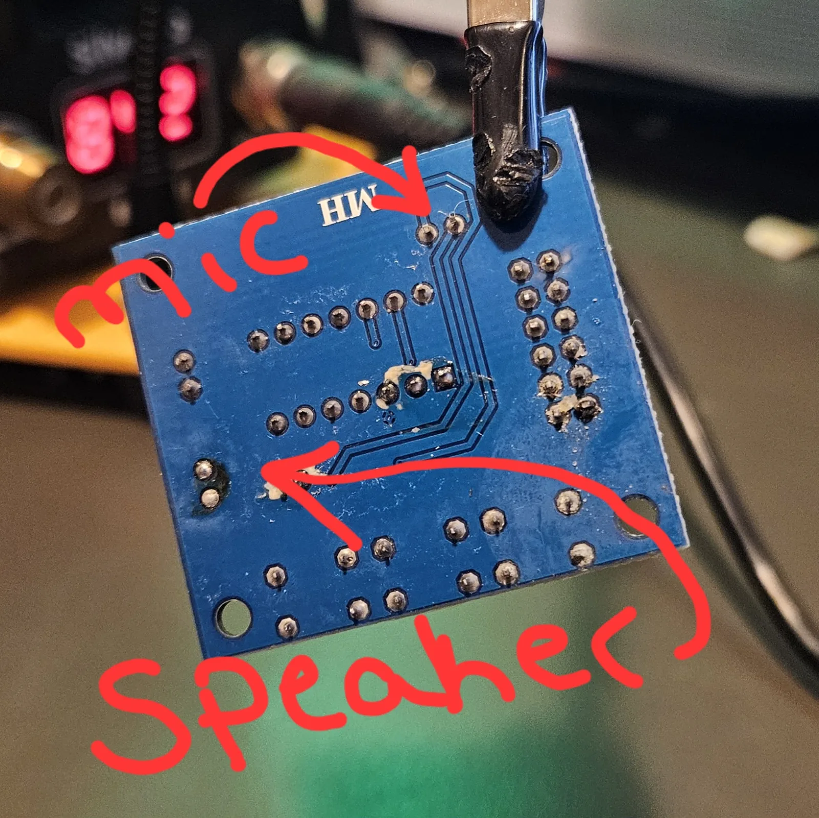 The ISD1820 module flipped to its back, held in a helping-hands clip. The bare blue underside shows rows of tinned through-hole pads and the silkscreen letters MH.