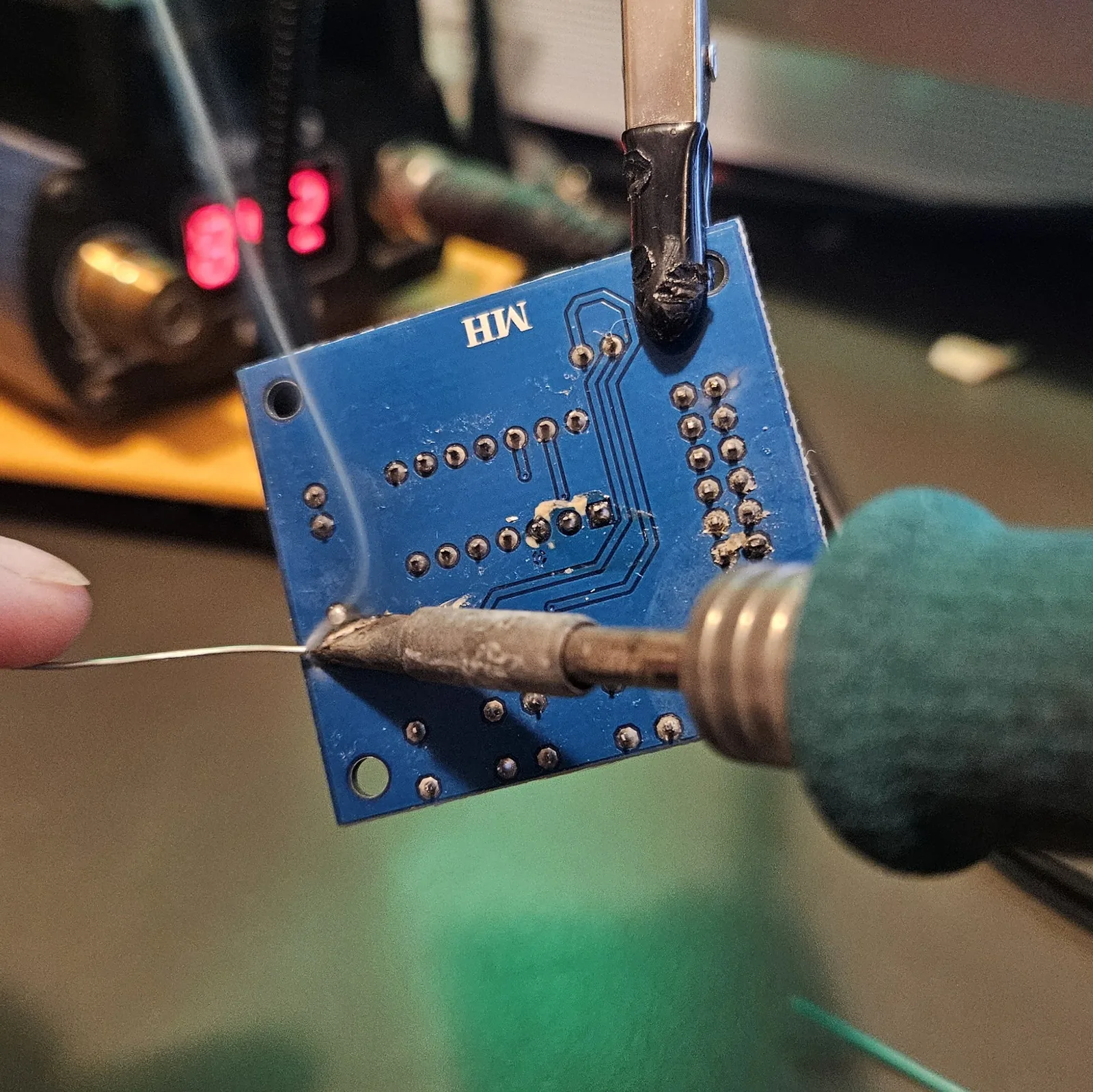 Desoldering the speaker connector: the board is clamped in a helping-hands clip while a soldering iron tip presses against a pad on the underside and a length of solder wire is fed straight onto the same joint, adding fresh solder rather than removing it. A soldering station with a red temperature display sits in the background.
