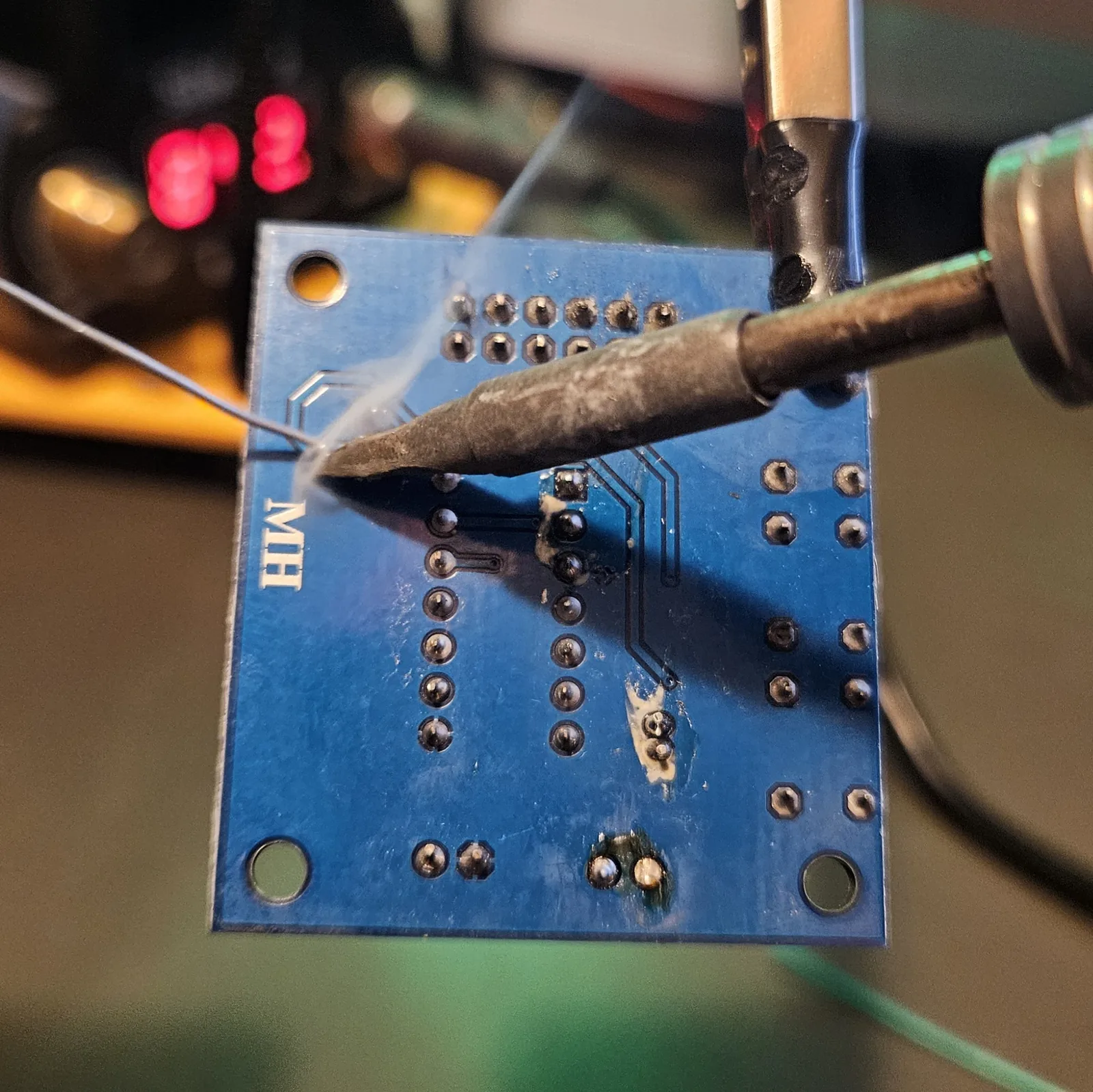 Desoldering the electret microphone with the same method: iron tip on the pad from the back of the board while solder wire is fed onto the joint, with the board held steady in the helping-hands clip.