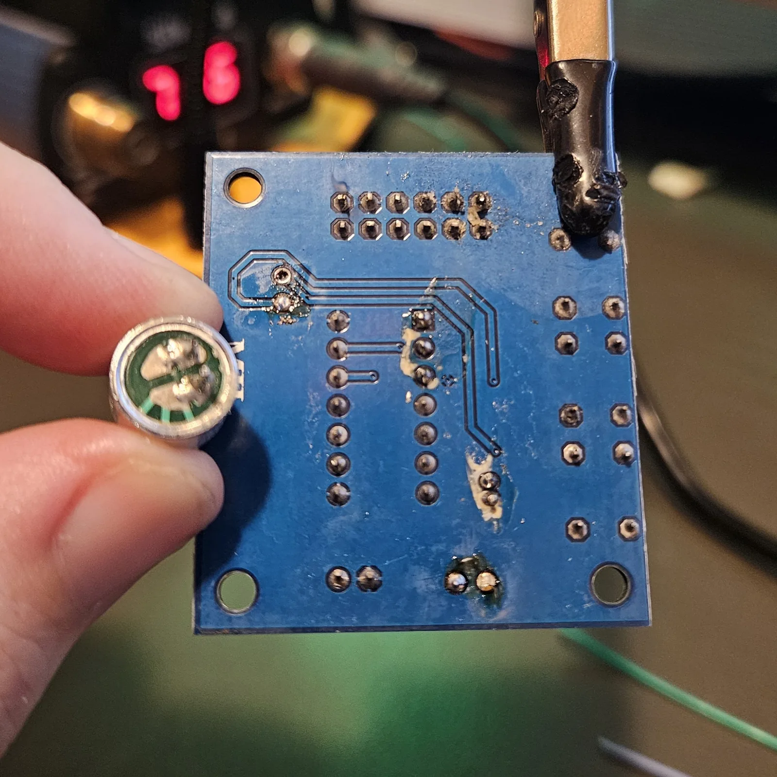 The freed electret microphone capsule held between finger and thumb, its green backing board and solder terminals visible, next to the stripped module with both the mic and speaker connector now removed.