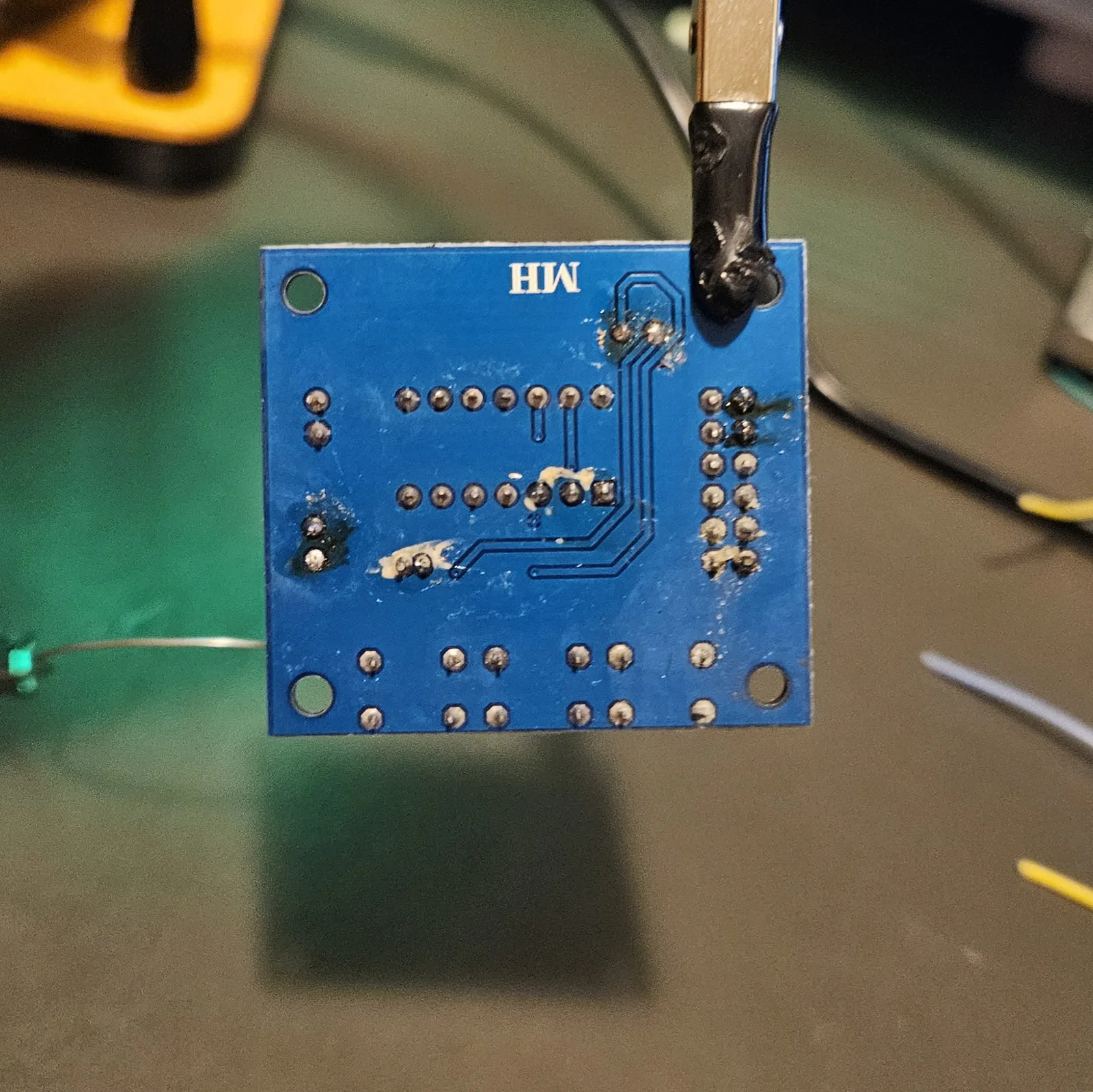 The stripped module held back-side-out in a helping-hands clip, its through-hole pads freshly tinned with small bright beads of solder ready to take wires.