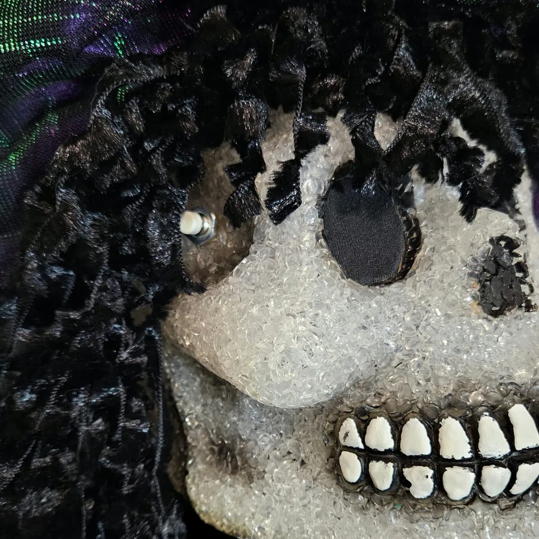 Close-up of the left temple: a small pushbutton with a white tip is mounted through the skull, half-buried in the black crushed-velvet ringlets. Below it the crystalline cheekbone catches the light, and the painted white teeth run along a black-lined jaw.