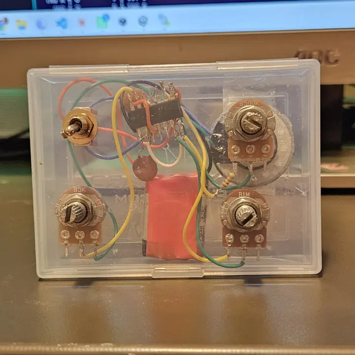 An Atari Punk Console built dead-bug style inside a translucent Carat playing-card box - an NE556 chip soldered legs-up in the center, four panel-mounted pots and a toggle, a salvaged toy speaker, a 9-volt battery, and hand-run wiring with no circuit board