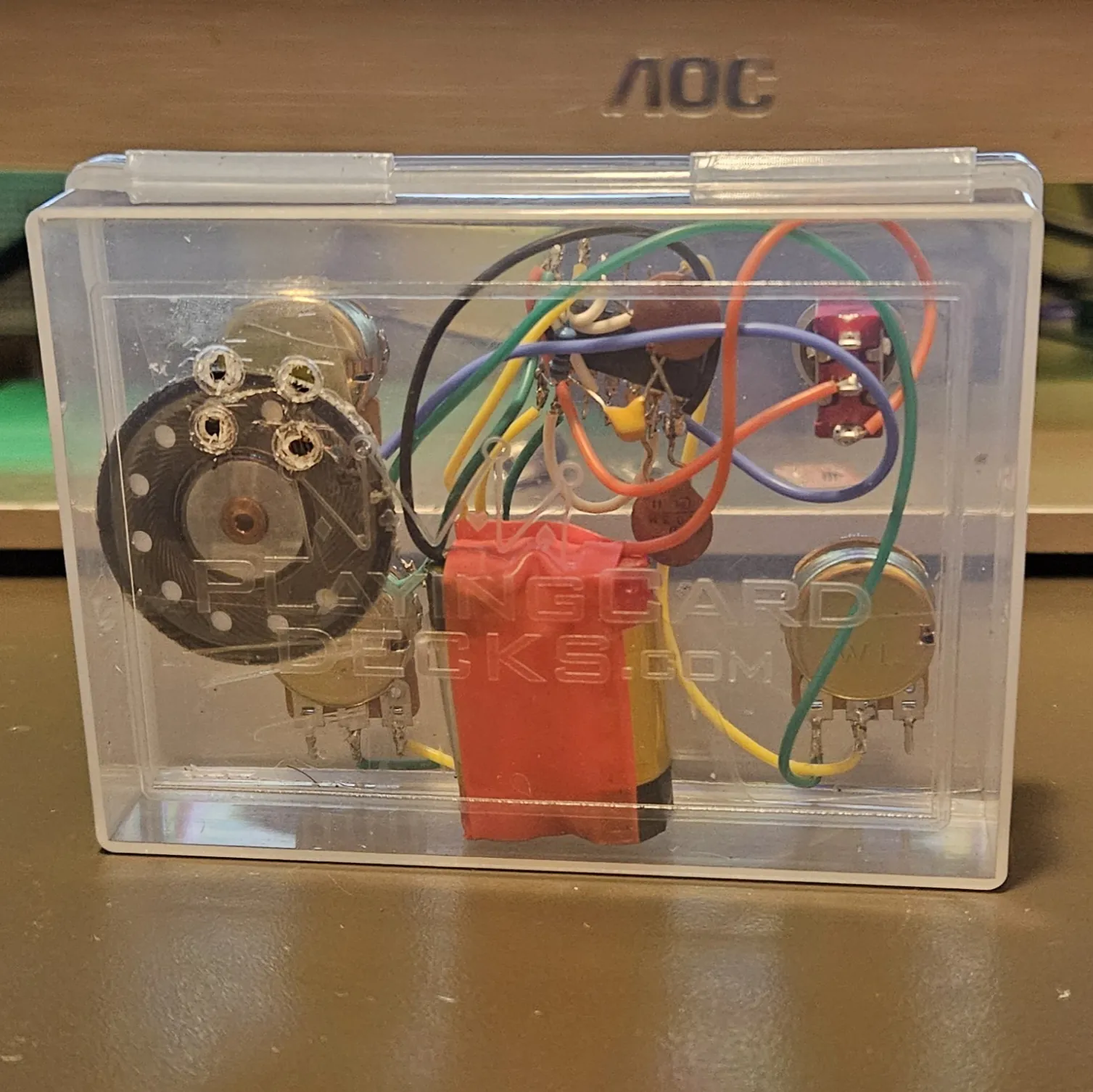 The closed box seen from the back, with PLAYINGCARDDECKS.COM embossed across the clear plastic. The whole circuit shows through it: the black speaker magnet at left, the gold pot bodies, the red toggle, the red-taped battery pack, and the nest of coloured wire behind them.