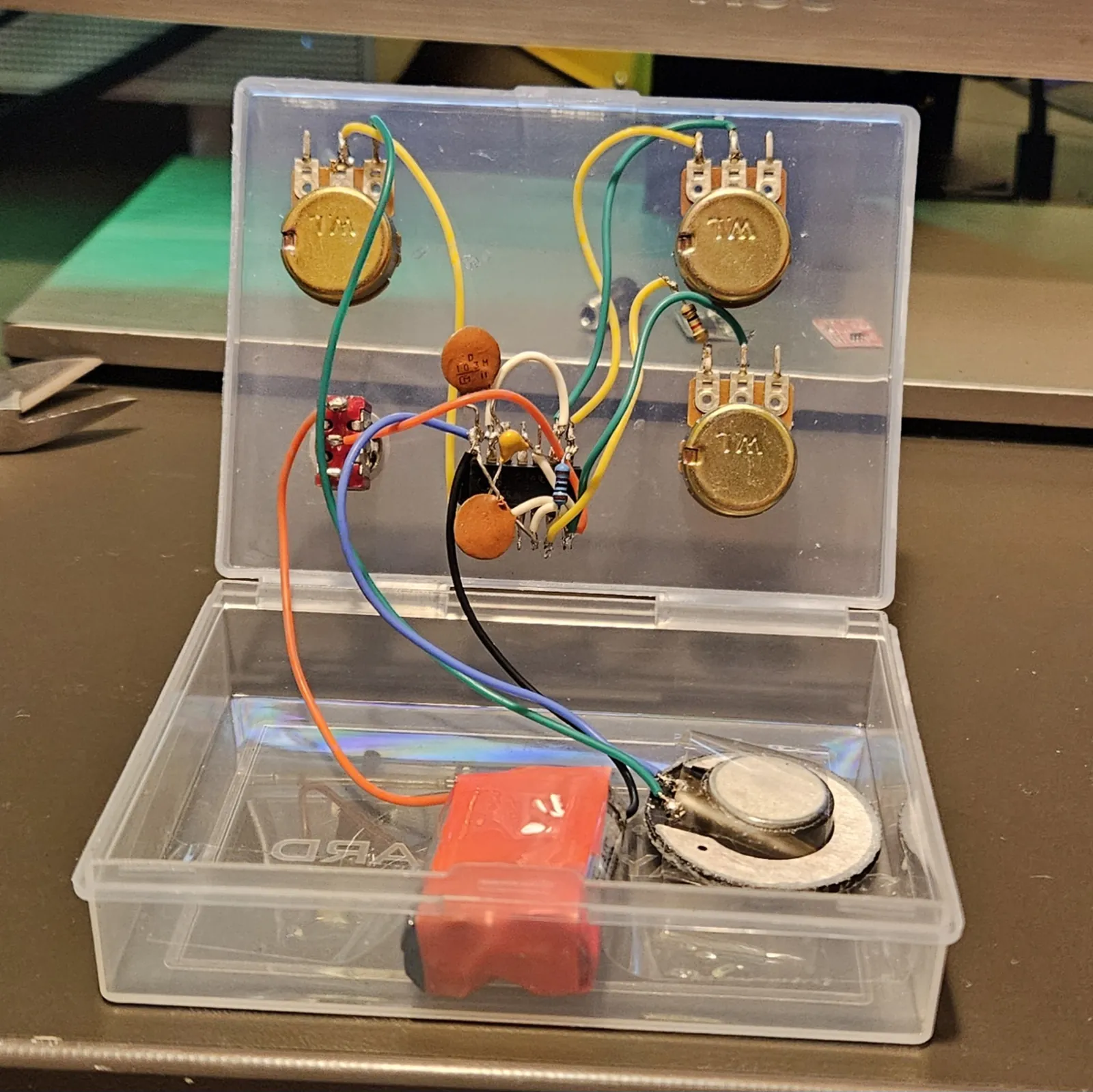 The card box open like a clamshell. Three gold W.L. potentiometers and a red toggle switch are mounted through the lid, with the NE556 soldered dead-bug in the middle of them - legs up, orange ceramic capacitors and a colour-banded resistor wired straight to its pins, and yellow, green, orange, blue, white and black hookup wire running out to each control. The lower half of the box holds the red-taped battery pack and the salvaged toy speaker.