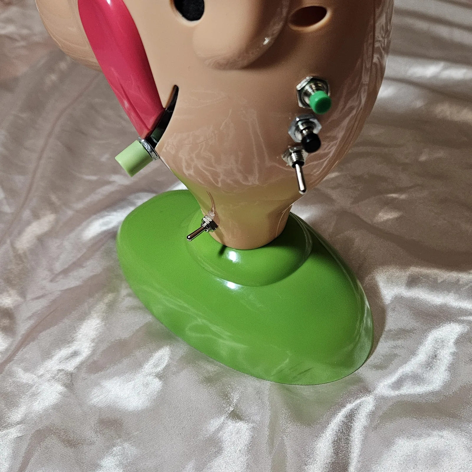 Tight view of the added controls on the head and neck: the green record button, the black play button below it, and the loop toggle with its silver lever pointing down, all in a vertical line on the side of the head. The green volume knob sits on the far side near the pink tongue, and the power switch is fitted low in the neck just above the green base.