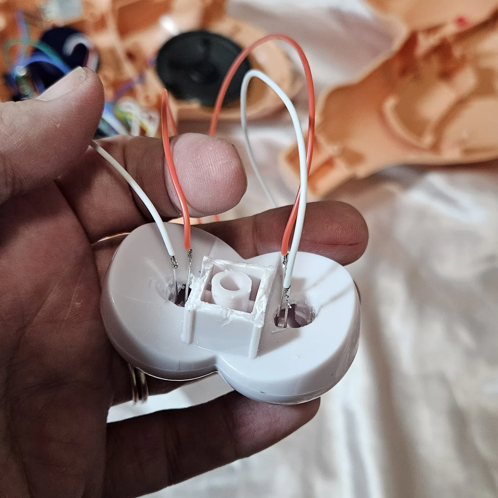 The eyeball assembly held in a hand, back side toward the camera: the white two-lobed plastic piece has a hole drilled through the back of each eye, with component leads pushed through and soldered to a red wire and a white wire on each side. White glue is packed around the central square post. Behind, out of focus, are the opened flesh-coloured shell, a black speaker and a tangle of coloured wire.