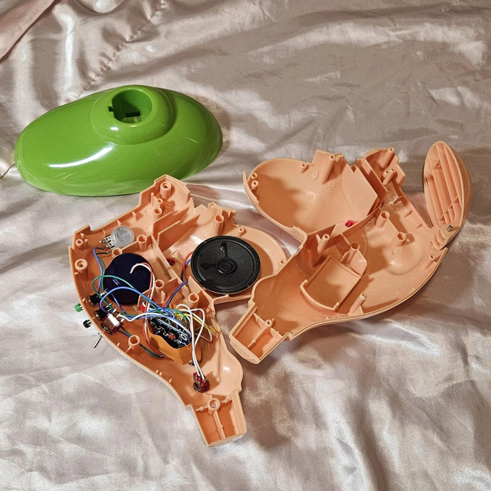 The guts: the left peach half-shell lying open and loaded, with the empty right half and the green base shell beside it. The blue ISD1820 recorder module sits low in the shell, its row of header pins fanned out to coloured wire; a round black speaker sits in the middle of the cavity, with a large blue capacitor and a small silver disc toward the top. Green, blue, white, yellow, orange and red hookup wire runs between them in a loose bundle. Along the shell rim are the mounted controls seen from inside - a green pushbutton, a black pushbutton, a silver potentiometer body and two mini toggle switches - and a small red part on twin white wires sits at the bottom.