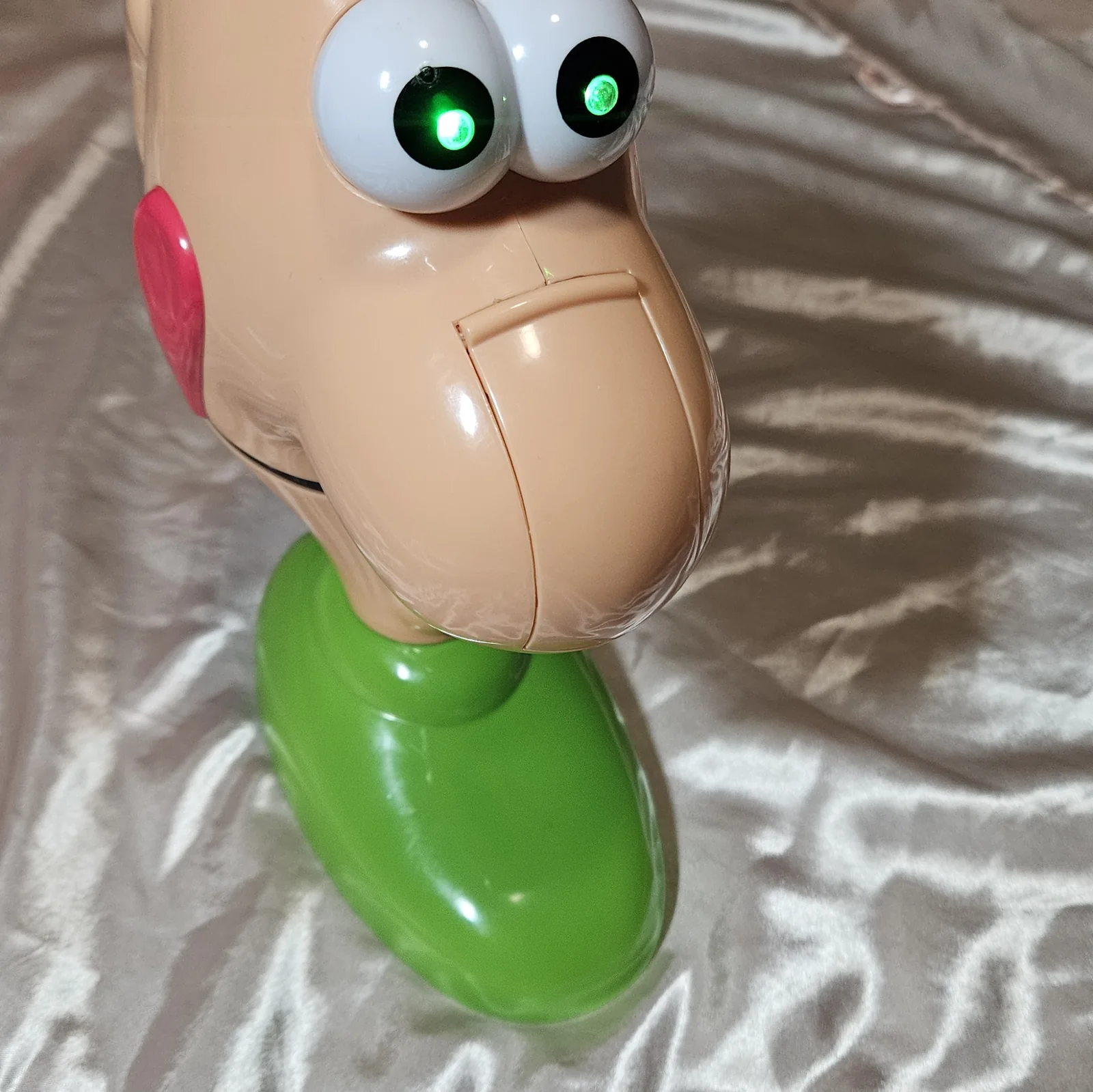 Close-up of the face: both white eyeballs fill the frame with a hard green point of light burning in the centre of each black pupil, one flaring brighter than the other. The huge moulded nose and the pink tongue at the corner of the mouth are lit only by the room, on shiny cream fabric.