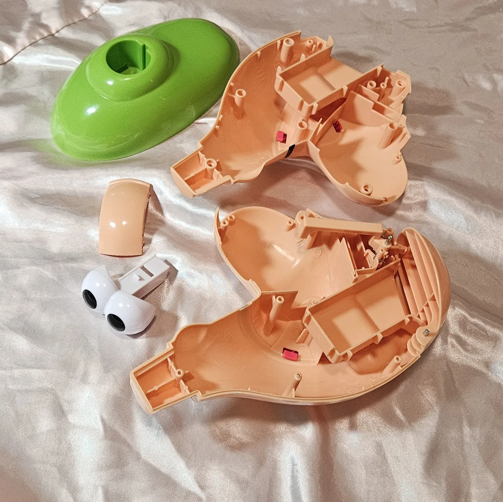 The toy stripped to bare plastic on cream fabric: the hollow green base shell at top left with its keyed socket, the two empty flesh-coloured halves of the head laid open to show ribbed moulding and small pink plastic tabs, a short curved flesh-coloured trim piece, and the white two-lobed eyeball assembly with its black pupils sitting next to a white plastic bracket. No electronics are fitted yet.