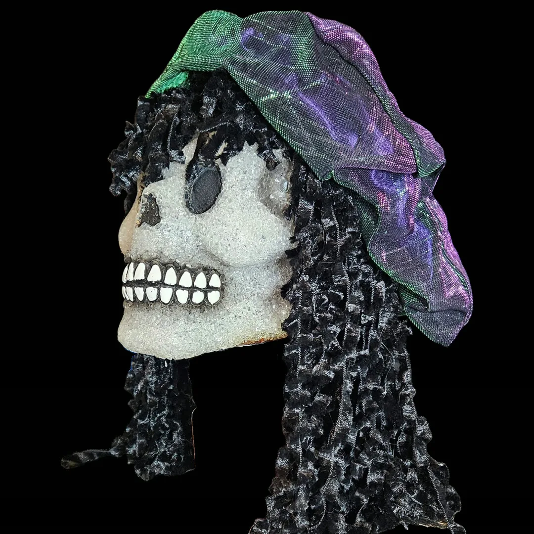 Full left profile against black: the skull's crystalline white face with a black teardrop eye socket and a grinning row of painted white teeth, framed by a long curtain of black ringlets that falls well below the jaw, with the iridescent cap draped over the crown.