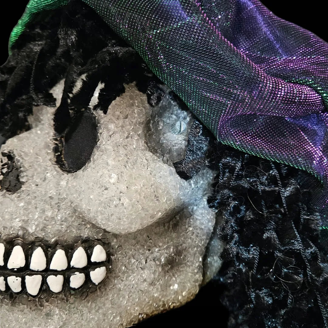 Close-up of the right temple: the pitch-bend potentiometer is mounted straight through the skull with no knob on it &mdash; just the bare metal shaft and its collar, sitting flush in the crystalline finish. The iridescent cap drapes overhead, black curls fall past the jaw, and the painted white teeth run along the lower left of the frame.