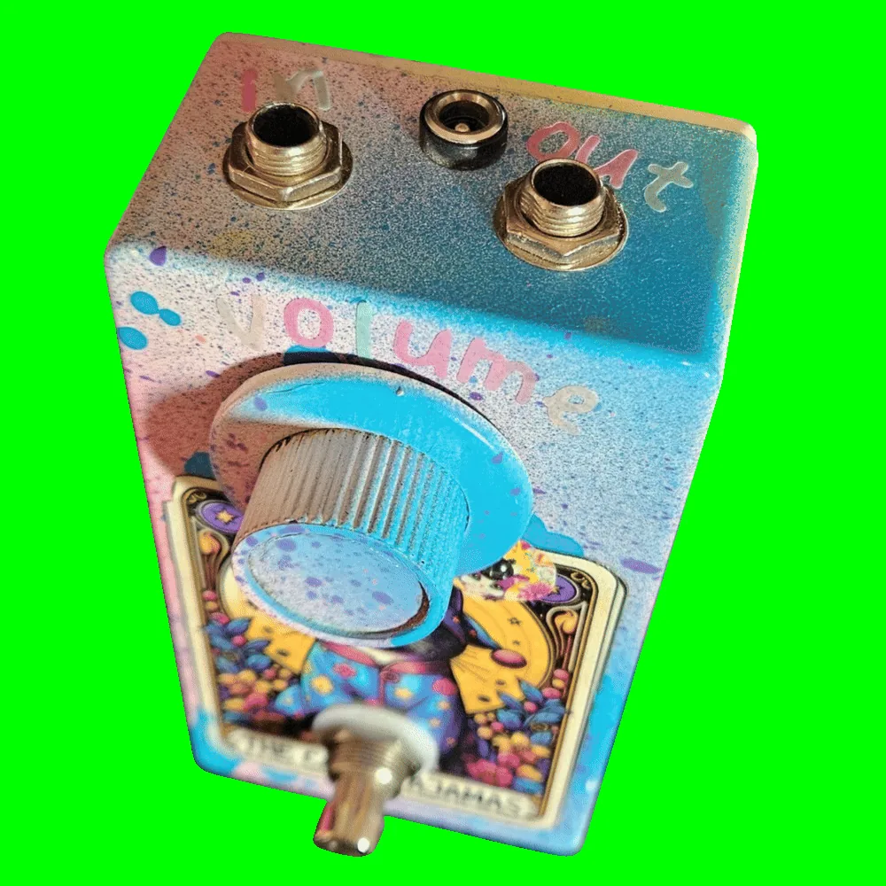 Top-down view of the enclosure: a 1/4-inch jack sits at each corner of the top edge, hand-lettered "in" on the left and "out" on the right, with a barrel DC power jack centred between them. Pastel letters spelling "volume" arc above the large light-blue knurled knob.