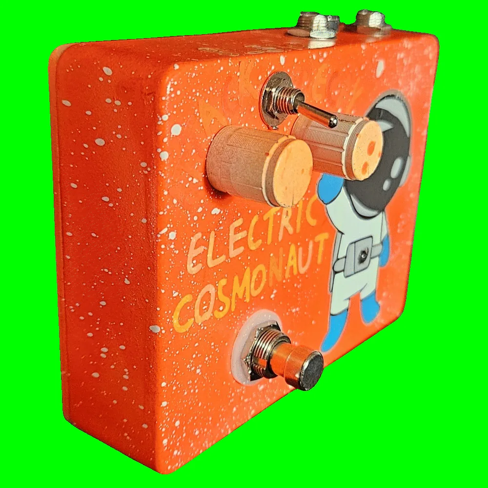 Left side of the pedal: an orange wall flecked with white speckle, showing the two cream knobs and the toggle switch standing out from the face and the footswitch protruding at the bottom. Hand-lettered "ELECTRIC COSMONAUT" runs across the front in cream and yellow beside the waving astronaut.