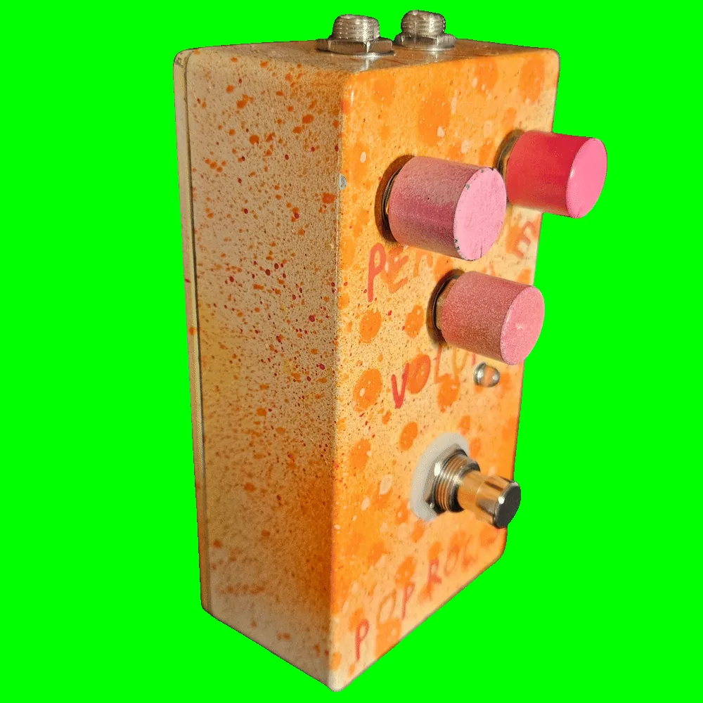 Left side of the pedal: a plain cream wall flecked with orange and red, showing the three pink barrel knobs standing off the face, a small LED beside the Volume knob, and the footswitch mounted on a white washer.