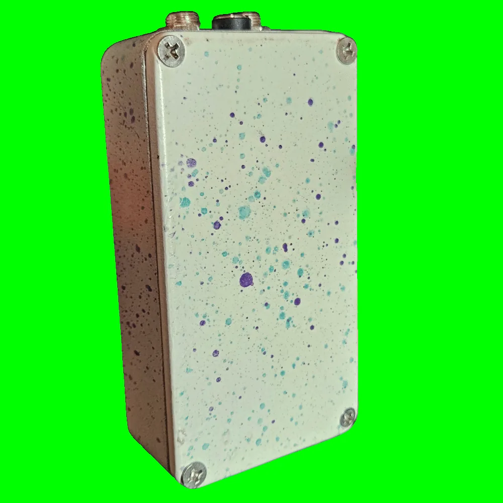Back of the pedal: a cream bottom plate scattered with teal and purple splatter dots, with a Phillips screw in each corner and no label. Two jacks and the DC socket show along the top edge.